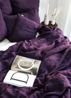 an open book sitting on top of a bed covered in purple sheets and pillows next to a candle