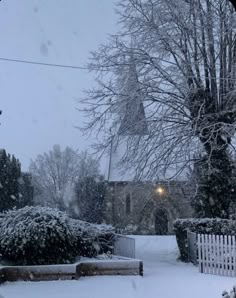 a snow covered yard with trees and bushes in the foreground is a church steeple