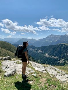 a woman standing on top of a grass covered hillside with mountains in the background and clouds in the sky