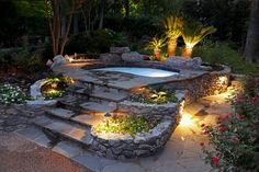 an outdoor hot tub surrounded by plants and rocks with lights on the steps leading up to it