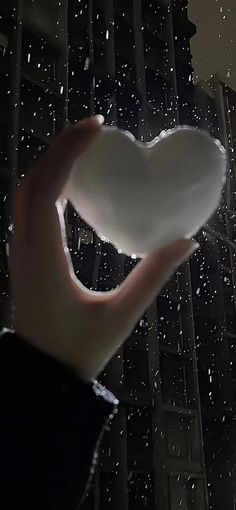 a hand holding a white heart shaped object in front of a rain soaked building at night