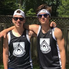 two young men standing next to each other in front of a wooden fence and trees