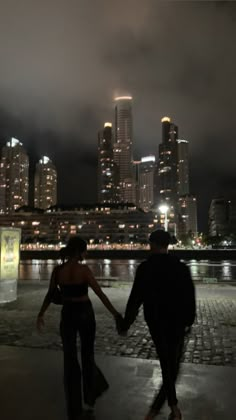 two people holding hands walking in front of a city at night with the lights on