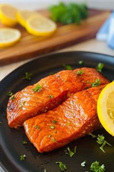 two pieces of salmon on a black plate with lemons and parsley next to it