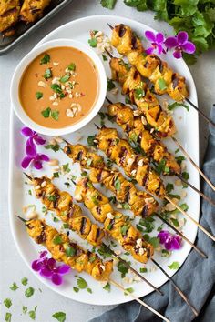 grilled chicken skewers with dipping sauce on a white plate next to purple flowers