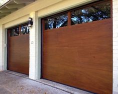 two brown garage doors in front of a white brick building with trees on the other side