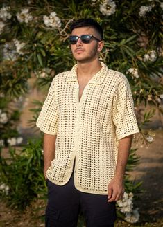 a man wearing sunglasses standing in front of some bushes and flowers with his hands in his pockets