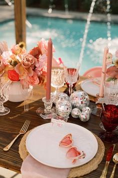 an outdoor table set with pink flowers, candles and plates near a pool in the background