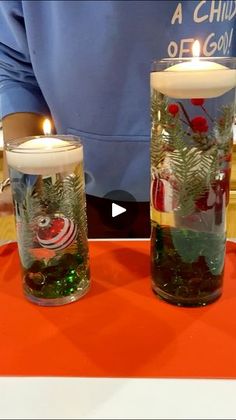 two glass vases filled with plants and candles on top of an orange tablecloth