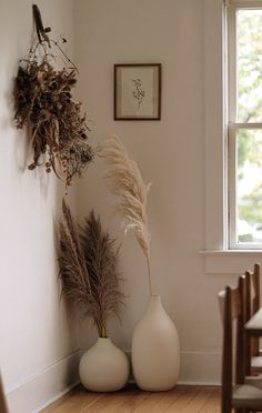 two white vases with dried plants in them on a wooden floor next to a window