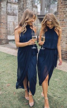 two women in blue dresses holding wine glasses and looking at each other while standing on the grass