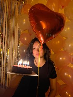 a woman holding a cake with candles in front of her face and balloons behind her