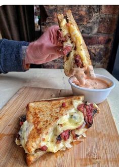 a person holding a sandwich over a wooden cutting board with a bowl of sauce in the background