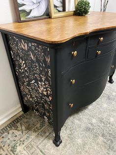 a black dresser with wooden top and gold knobs on the drawers is next to a framed painting