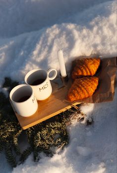 three coffee mugs and two croissants on a tray in the snow