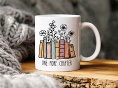 a white coffee mug with flowers and books on it sitting on a wooden table next to a gray sweater