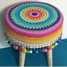 a multicolored crocheted stool with wooden legs and pom poms