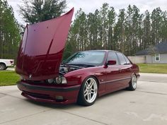 a red car with its hood open in front of a driveway and trees behind it