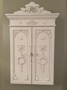 a white cabinet with ornate carvings on the front and sides, against a gray wall