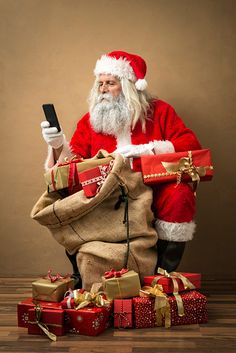 a man dressed as santa claus holding a cell phone in his hand while sitting on presents