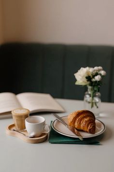 a croissant on a plate next to a cup of coffee and an open book