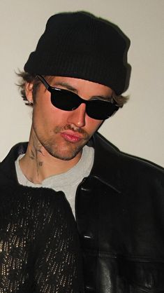 a man wearing sunglasses and a hat looks at his cell phone while standing in front of a white wall