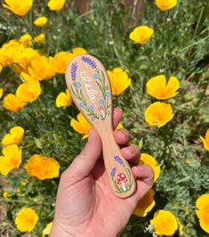 a hand holding a wooden spoon in front of yellow flowers