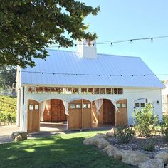 a white barn with wooden doors and windows on the front side is surrounded by greenery