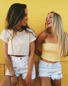 two young women standing next to each other near a yellow wall and one is holding her purse