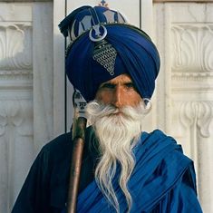 a man with a long white beard wearing a blue turban and holding a stick