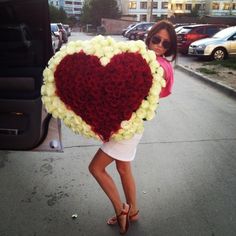 a woman holding a heart shaped arrangement of flowers in front of a car parked on the street