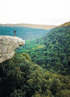 a person standing on the edge of a cliff looking down at some trees and mountains