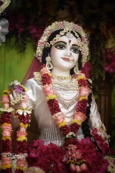 a statue with flowers around it in front of a green wall and some pink flowers