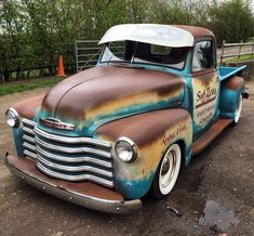 an old truck with rusted paint parked on the side of a dirt road in front of a fence