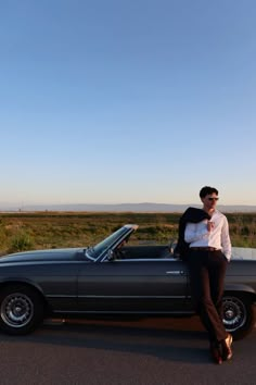 a man leaning on the hood of a black convertible car in front of a field