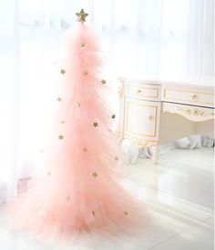 a pink christmas tree with gold stars on it in front of a white dresser and window