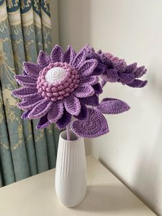 a crocheted purple flower in a white vase on a table next to curtains