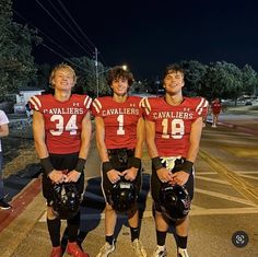 three young men in football uniforms standing on the side walk at night with helmets around their necks