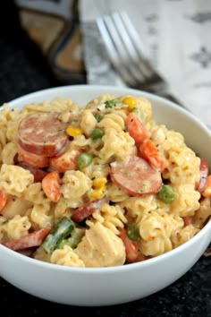 a white bowl filled with pasta and vegetables next to a silver fork on top of a table