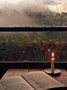 a candle sitting on top of an open book next to a window with raindrops