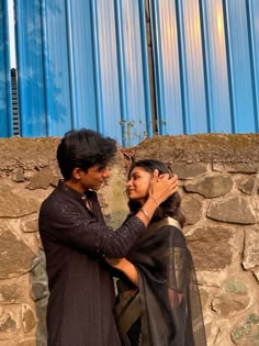 a man and woman standing next to each other near a stone wall with a blue building in the background