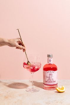 a person is holding a stick near a glass with a pink liquid in it and a bottle next to it