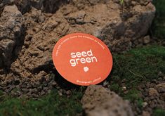 a red sign that says seed green sitting on the ground next to some rocks and grass