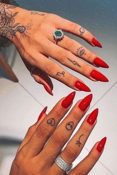 two hands with red nail polish and tattoos on their fingers, one holding an emerald ring