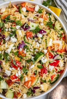 a bowl filled with pasta salad and topped with feta cheese, tomatoes, cucumbers, red onion, parsley and lemon wedges