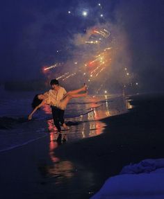 two people dancing on the beach at night with fireworks in the sky above them and water below