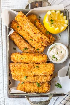 fried fish sticks with lemon wedges and tartar sauce in a tray on a white wooden table