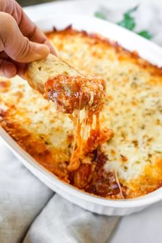 a hand holding a piece of pizza with cheese and sauce on it in a white casserole dish