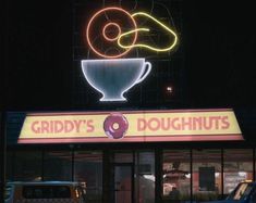 there is a doughnut shop at night with neon lights on the building and cars parked outside