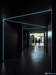 two people are walking through an empty hallway with blue lights on the ceiling and black walls